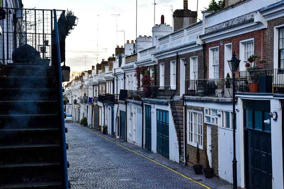 A narrow residential street in Holland Park featuring a row of terraced houses with brick and white-painted facades, some with decorative cornices and window frames. The street is paved with cobblestones and lined with black metal railings, flower pots, and small potted plants on the balconies. A black metal lamppost is visible on the right side of the image. On the left side, a staircase with a metal railing and steps leading up to a higher level is partially visible. The sky in the background shows a sunset or early evening lighting, casting a soft glow over the scene. This setting, typical of house relocation services, highlights the tight urban environment where careful furniture transport and packing are essential, as managed by companies like Holland Park Movers for stress-free home removals and moving logistics.