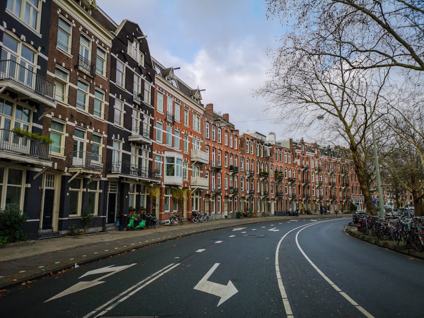 A row of traditional Victorian-style terraced houses on Holland Park Road in W8, London, featuring brick facades, bay windows, small balconies with wrought iron railings, and multiple stories. The street is lined with leafless trees, and the pavement is occupied by parked bicycles and scooters. A building entrance with steps and a small entrance porch is visible at the street level. The road curves to the right, marked with white double lines and directional arrows, with parked cars along the curb. The sky is partly cloudy, and the scene suggests an urban residential area. Holland Park Movers may use this location for furniture transport or home relocation services, as part of their house removals process involving loading or unloading in a typical residential street setting.