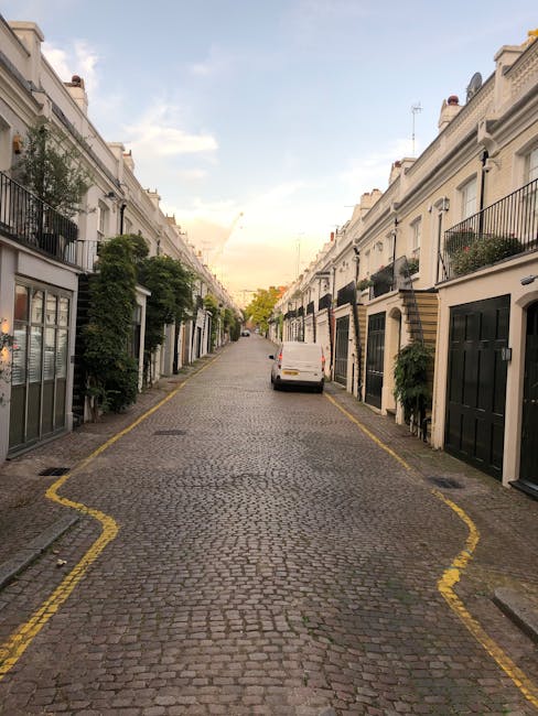 A narrow residential street with a cobblestone surface, flanked by white terraced houses featuring black and beige doors and iron railings on upper balconies. Greenery, including potted plants and small trees, is visible along the front of the buildings. In the middle of the street, a white van is parked facing away, with its rear doors partially accessible for loading or unloading. The street lines are marked with yellow curb paint indicating parking restrictions or loading zones. Overhead, the sky is partly cloudy with soft sunlight illuminating the scene, suggesting a late afternoon time. Holland Park Movers is depicted as potentially involved in the home relocation process, working within this tight urban environment, possibly during packing, furniture transport, or loading activities, with vehicles and materials suited for steps in a house removal or moving service.