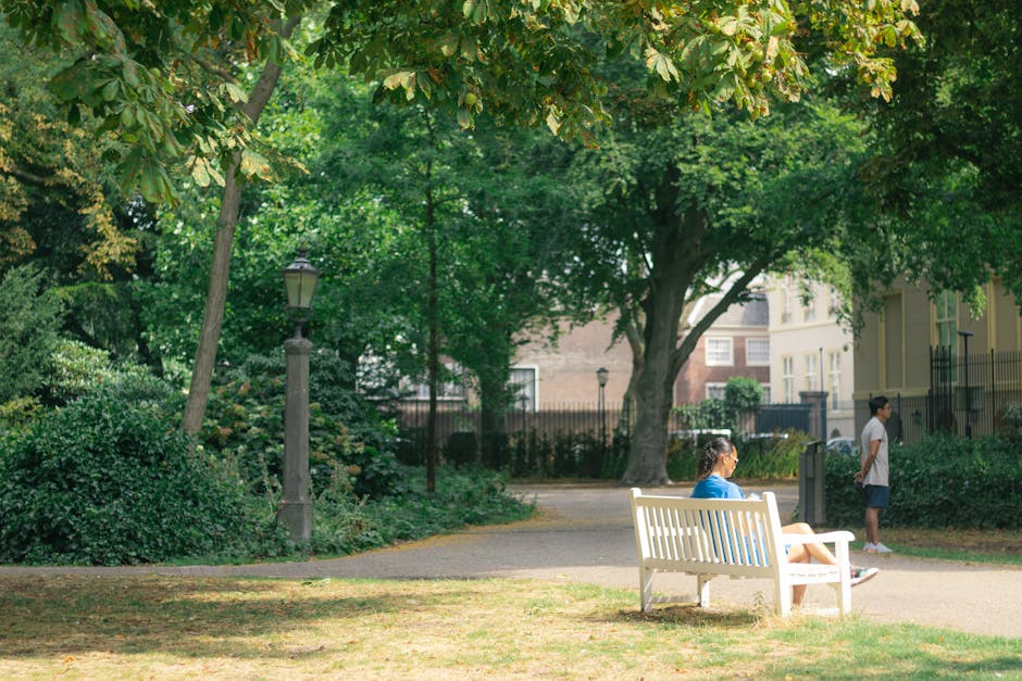 A woman sitting on a white park bench in a green outdoor setting, surrounded by lush trees with dense foliage and a lamppost nearby. In the background, two young individuals, a girl in a blue shirt and shorts and a boy in a white shirt and dark shorts, are standing on a paved pathway near residential buildings with a fence. The scene appears to be in a quiet urban park, with dappled sunlight filtering through the trees. This outdoor environment could be part of a home relocation or community moving process involving transportation or packing, as implied by Holland Park Movers' services for house removals near Notting Hill Gate. The overall scene emphasizes the natural surroundings and casual outdoor activity, suitable for illustrating the logistics of home relocation in a peaceful neighborhood setting.