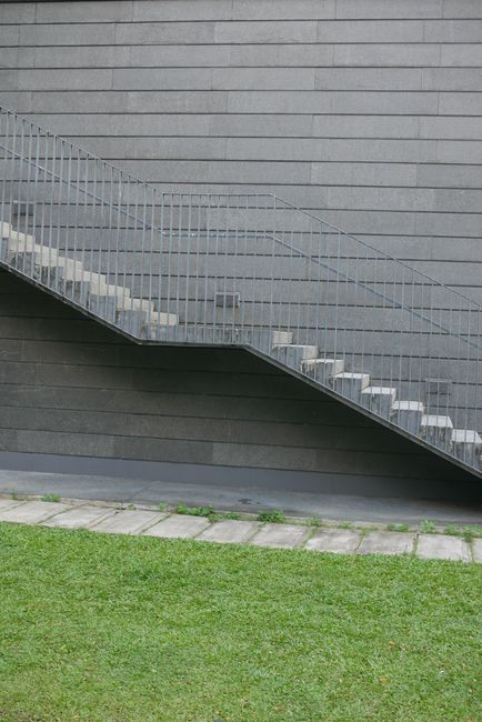 A modern exterior staircase with metal railings and white steps, positioned against a dark grey building wall with horizontal siding. The staircase is connected to a narrow outdoor area, with concrete paving stones and green grass in the foreground. The image depicts a location with limited access for moving large items, reflecting potential staircase access problems during a home relocation. This scene relates to house removals and furniture transport, where careful planning is required to navigate tight staircases, as typically managed by Holland Park Movers in moving and packing services.
