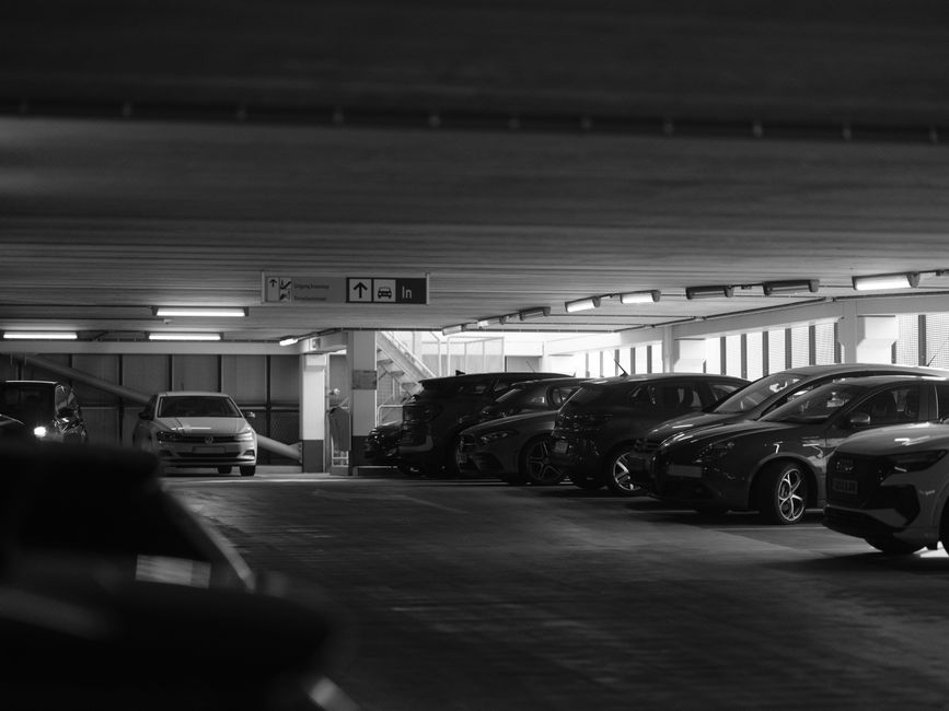Black and white photograph of an indoor parking garage with multiple parked cars arranged in two rows, including sedans and hatchbacks, positioned near the building's back wall and closer to the camera in the foreground. The garage features a low ceiling with visible wooden beams, and overhead fluorescent lighting illuminates the space. A sign hanging from the ceiling directs vehicles and pedestrians, with arrows indicating directions for entering or exiting the parking area, relevant to the context of Holland Park house removals and transport logistics. The environment appears functional and well-organized, typical of a vehicle storage and loading zone used during home relocation or furniture transport processes handled by Holland Park Movers.